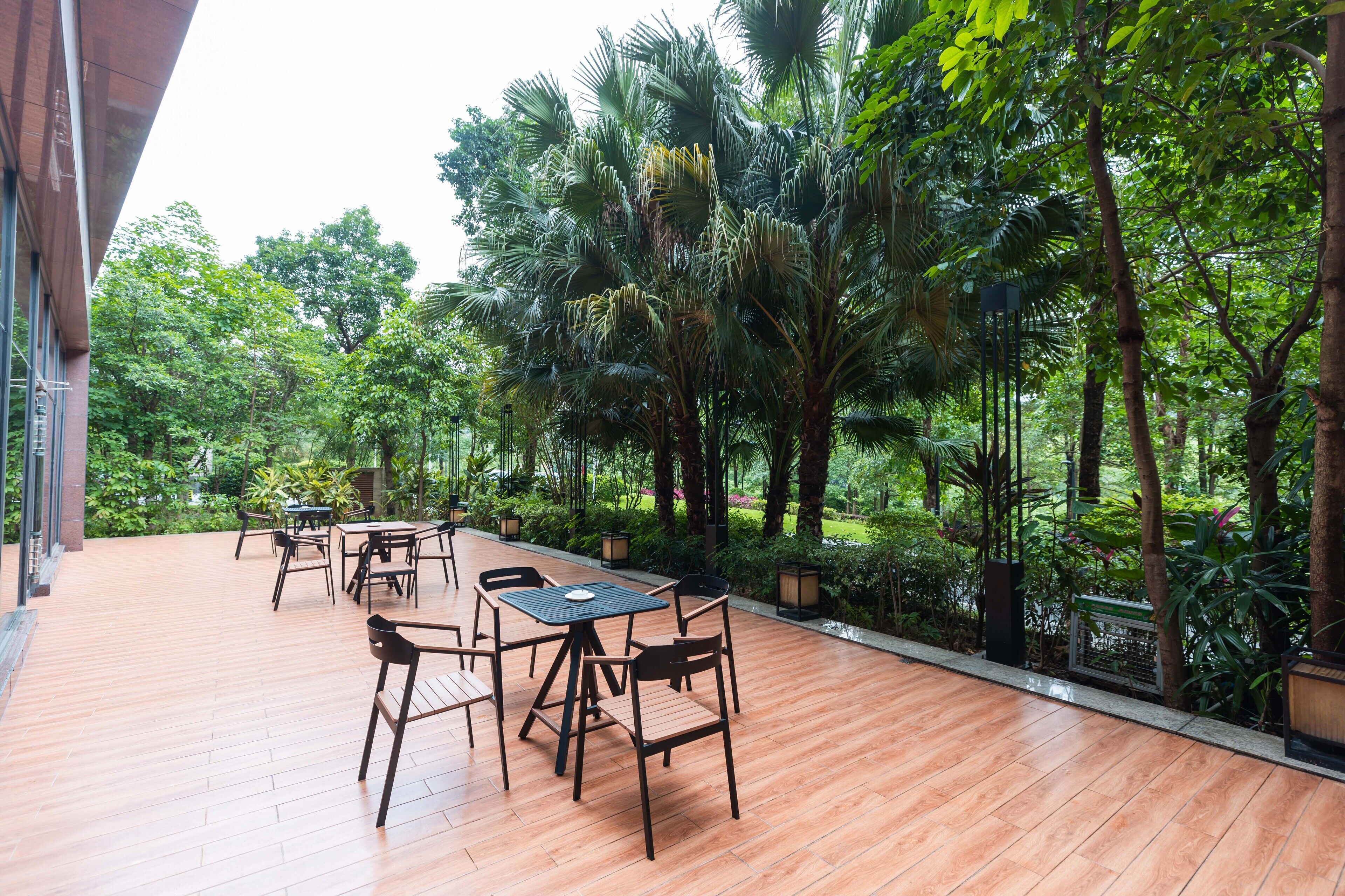 An outdoor patio with wooden tables and chairs, surrounded by lush greenery and tall trees.