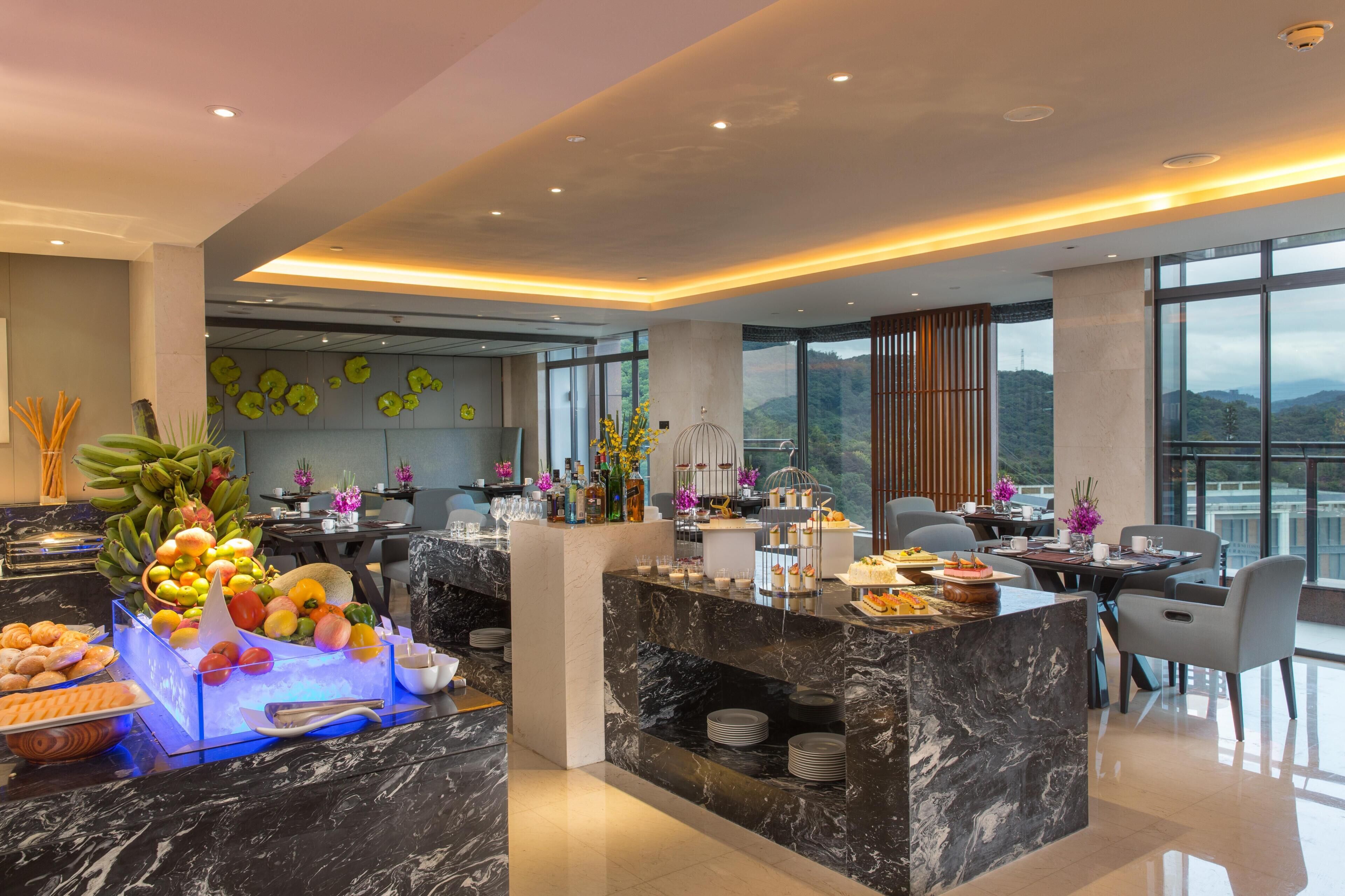 Un hall d’hôtel moderne avec un comptoir d’accueil en marbre, un présentoir de fruits et un espace de restauration offrant une vue sur les montagnes.