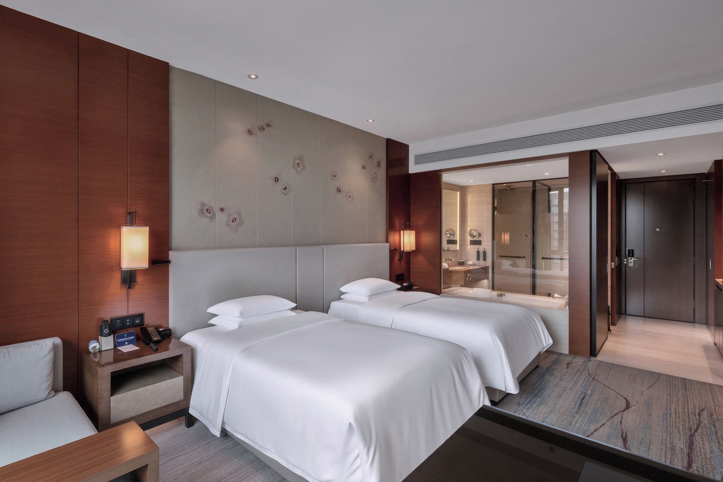 A hotel room with two beds, a bedside table with a phone and a lamp, a mirror, and a bathroom with a glass door.