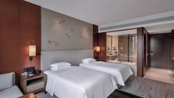 A hotel room with two beds, a bedside table with a phone and a lamp, a mirror, and a bathroom with a glass door.