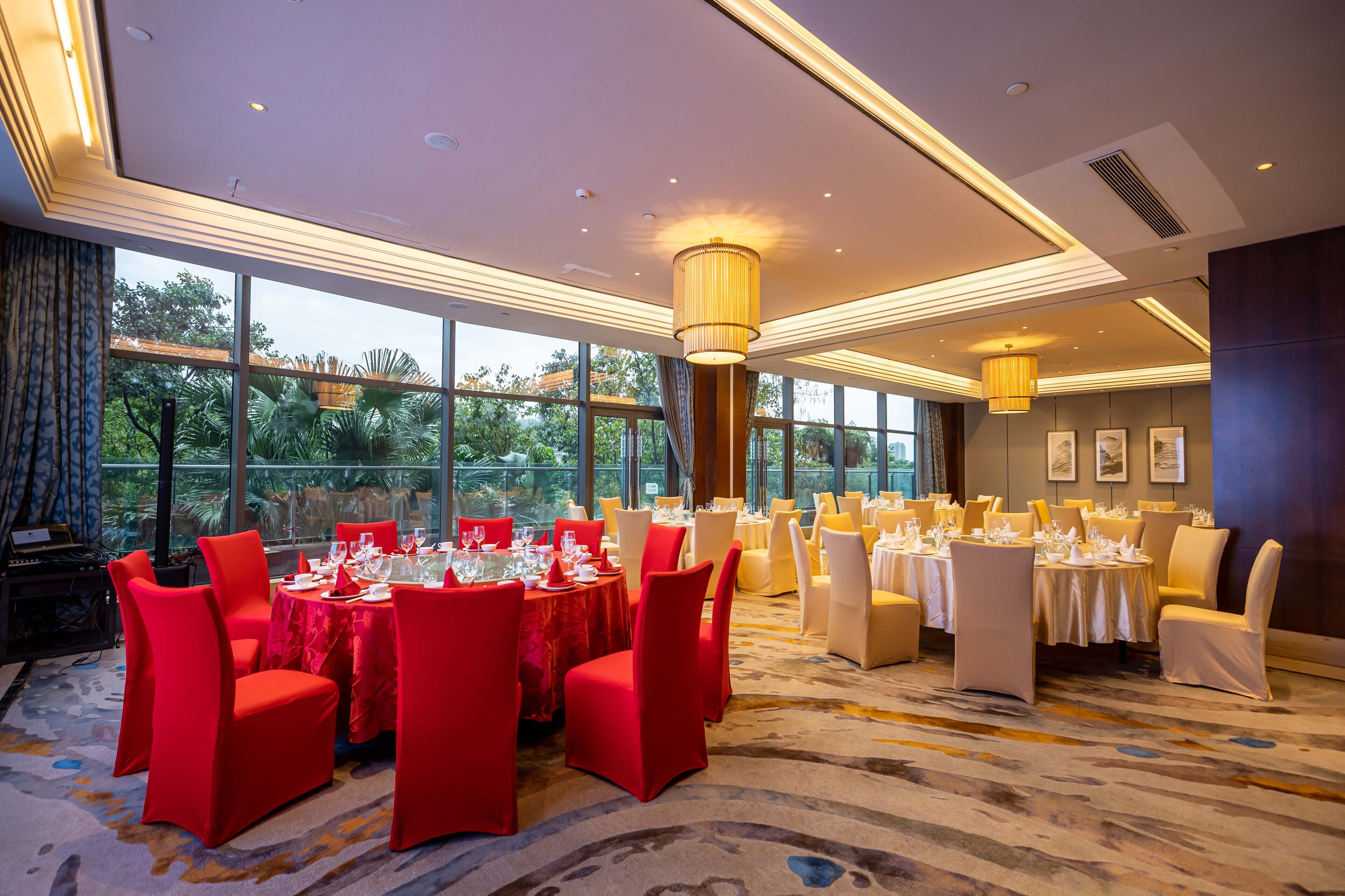 A banquet hall with round tables, red chairs, and a view of greenery outside.