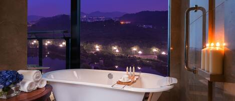 Ein modernes Badezimmer mit einer freistehenden Badewanne, einem kleinen Tisch mit Handtüchern und Pflegeprodukten und Blick auf eine Stadtlandschaft im Abendlicht.
