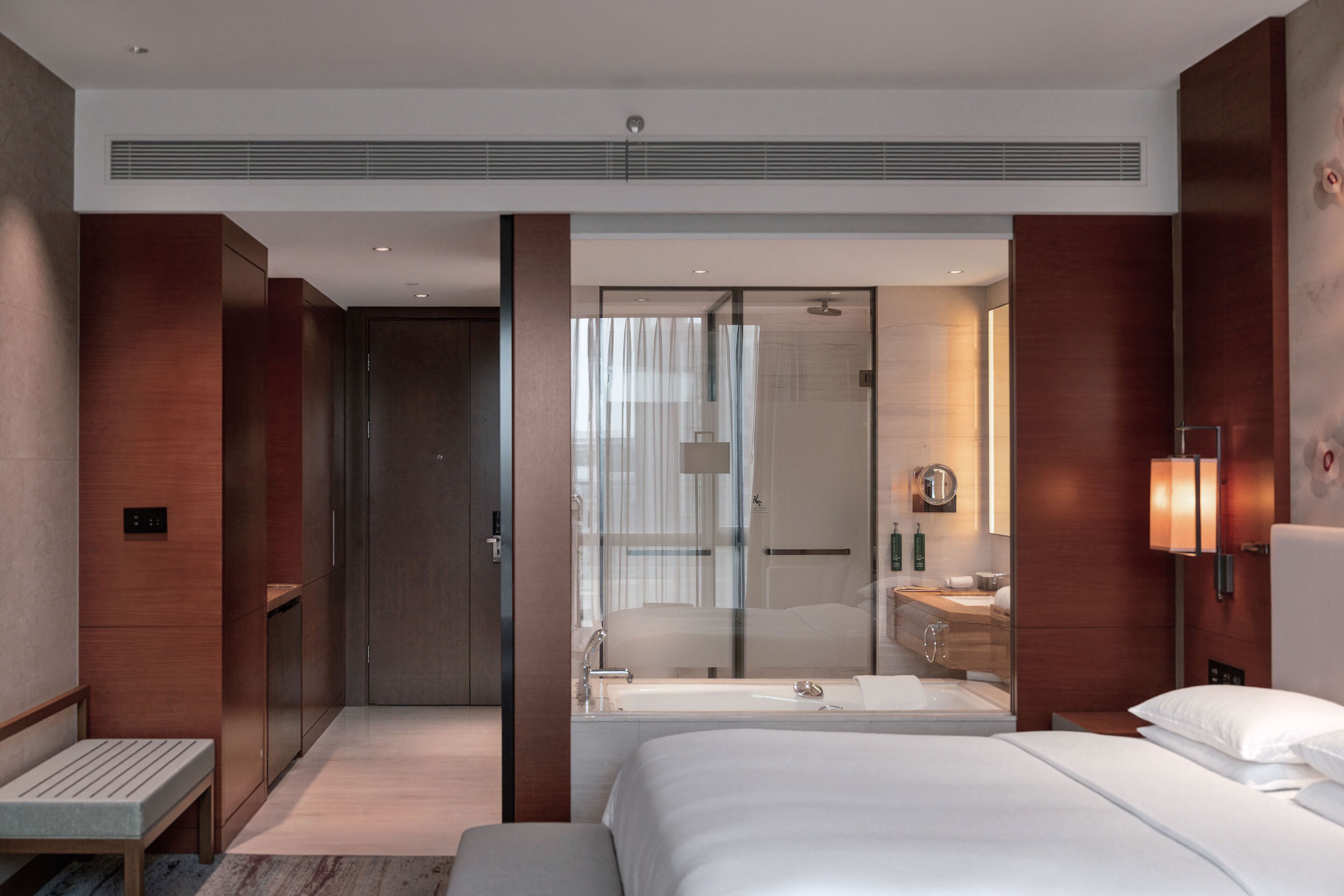 Une chambre d’hôtel moderne dotée d’un grand lit, d’une banquette, d’un dressing et d’une salle de bain avec un miroir et un lavabo.