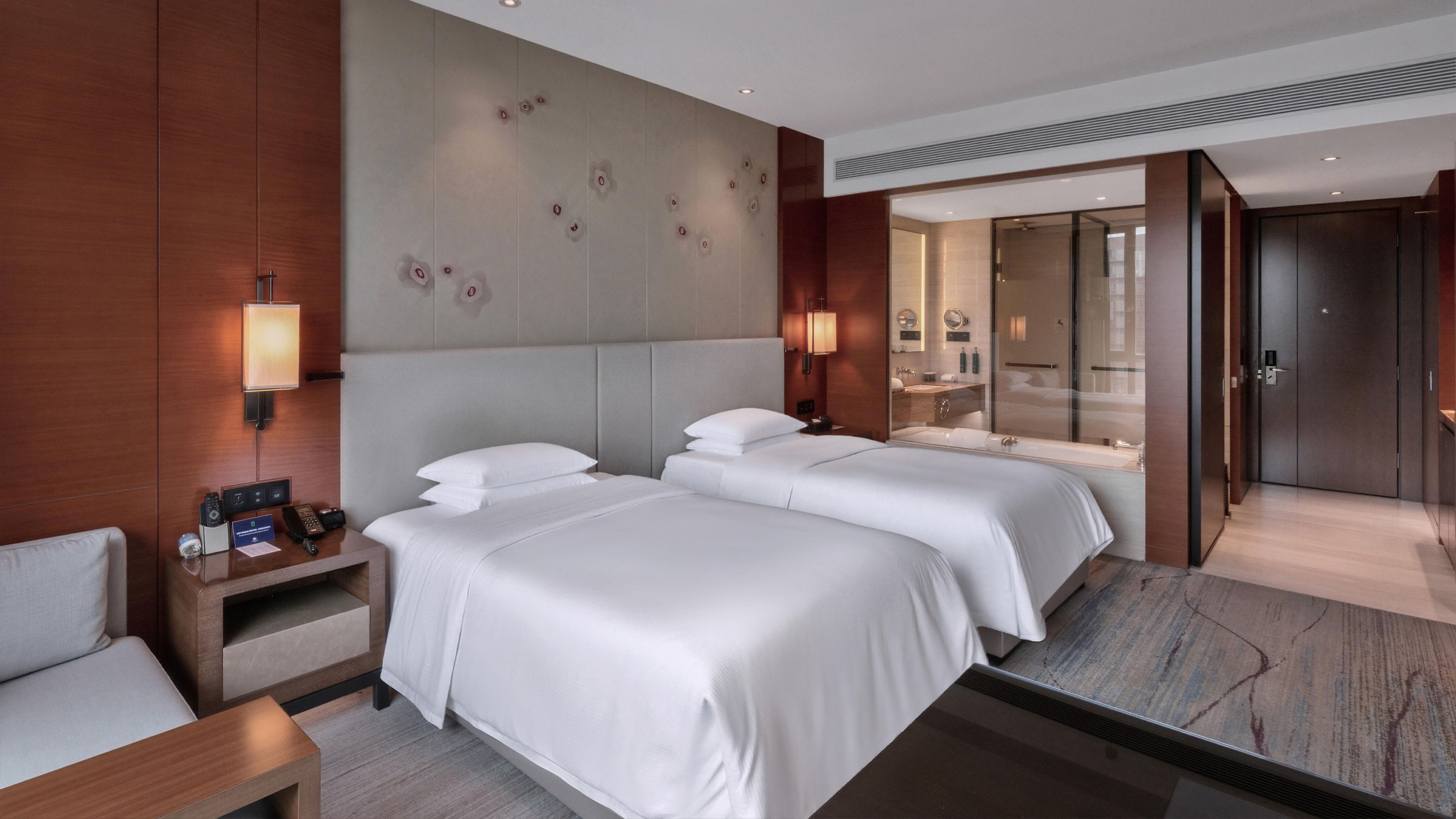 A hotel room with two beds, a desk, a chair, a bathroom with a shower, and a door leading to another room.