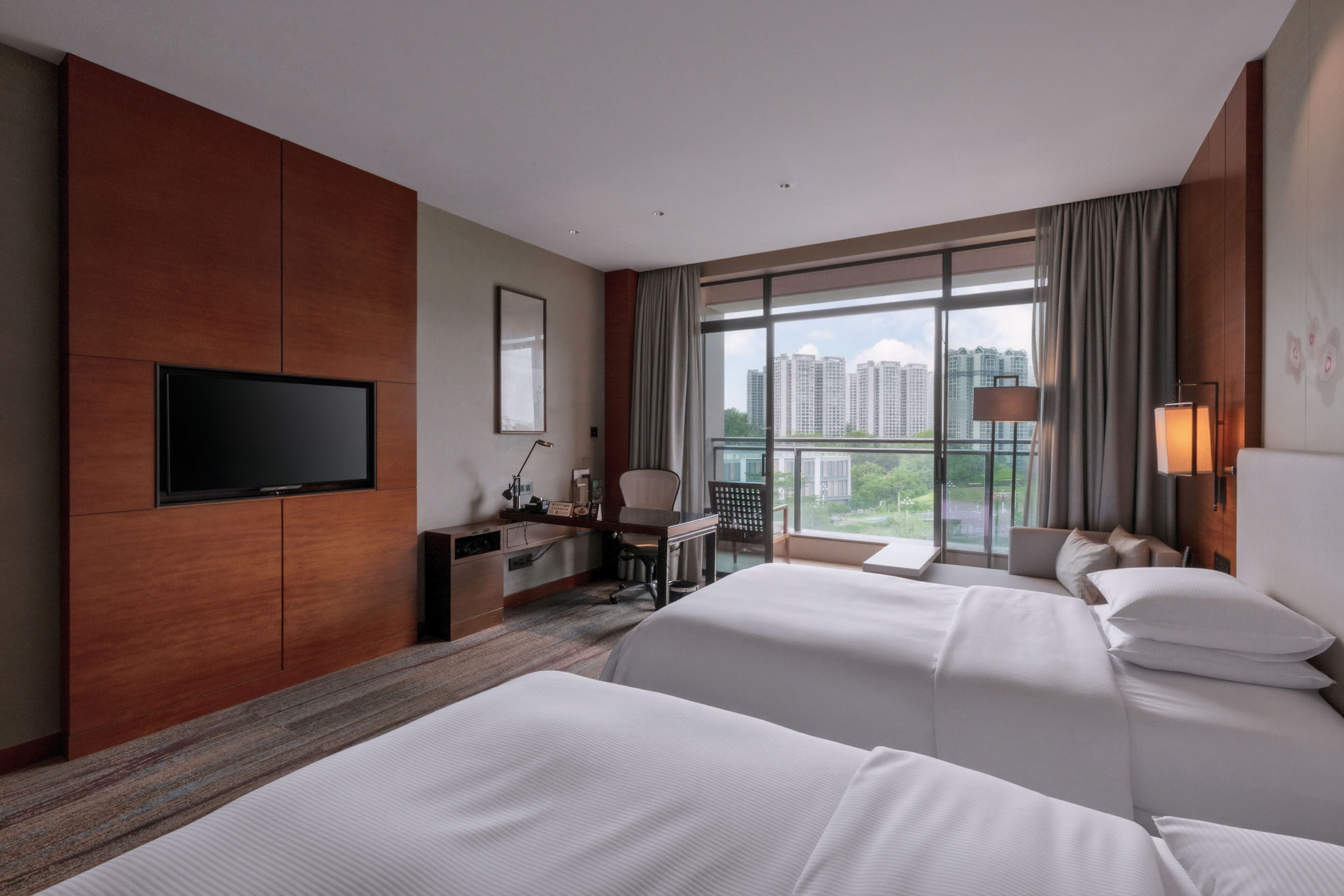 A hotel room with two beds, a TV, a desk, and a large window with a city view.