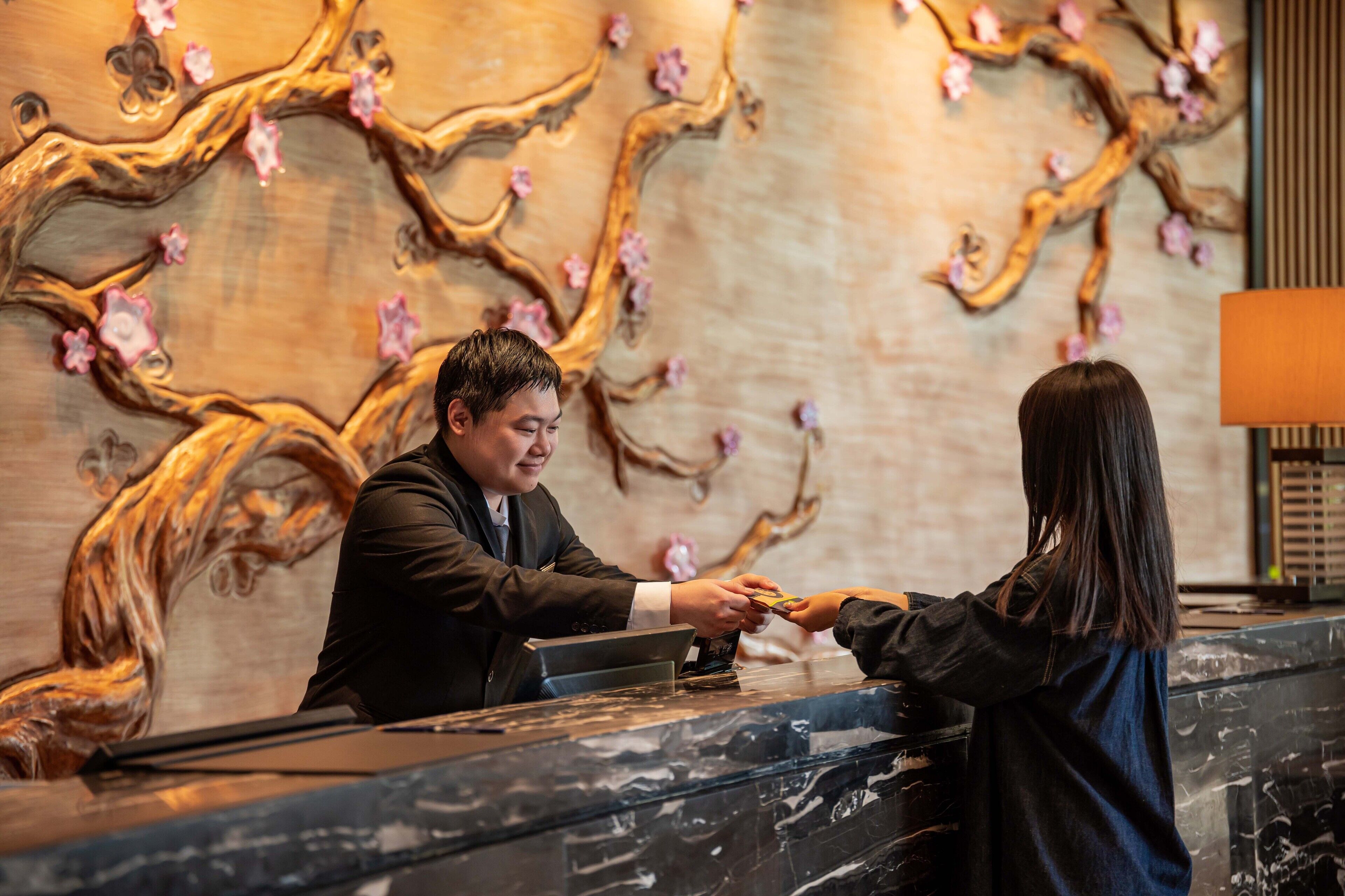 A reception desk with a marble counter, a man in a suit handing over a card to a woman, and a wall with a tree branch design and pink flowers.