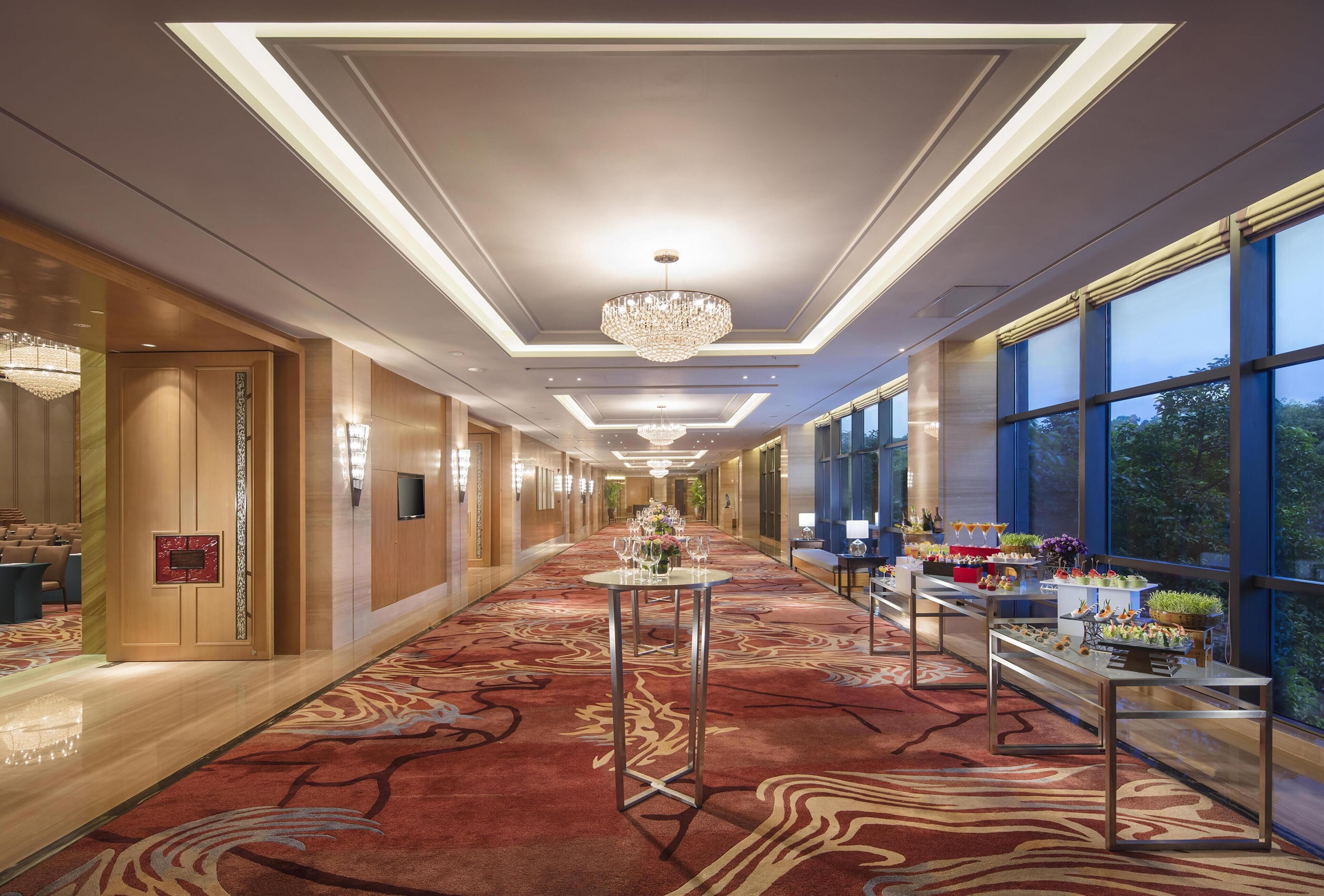 Un pasillo de hotel con alfombra estampada, un candelabro y mesas adornadas con flores.