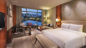 Quarto de hotel com uma cama grande, uma mesa com cadeira, uma televisão e vista para a paisagem urbana através de amplas janelas.