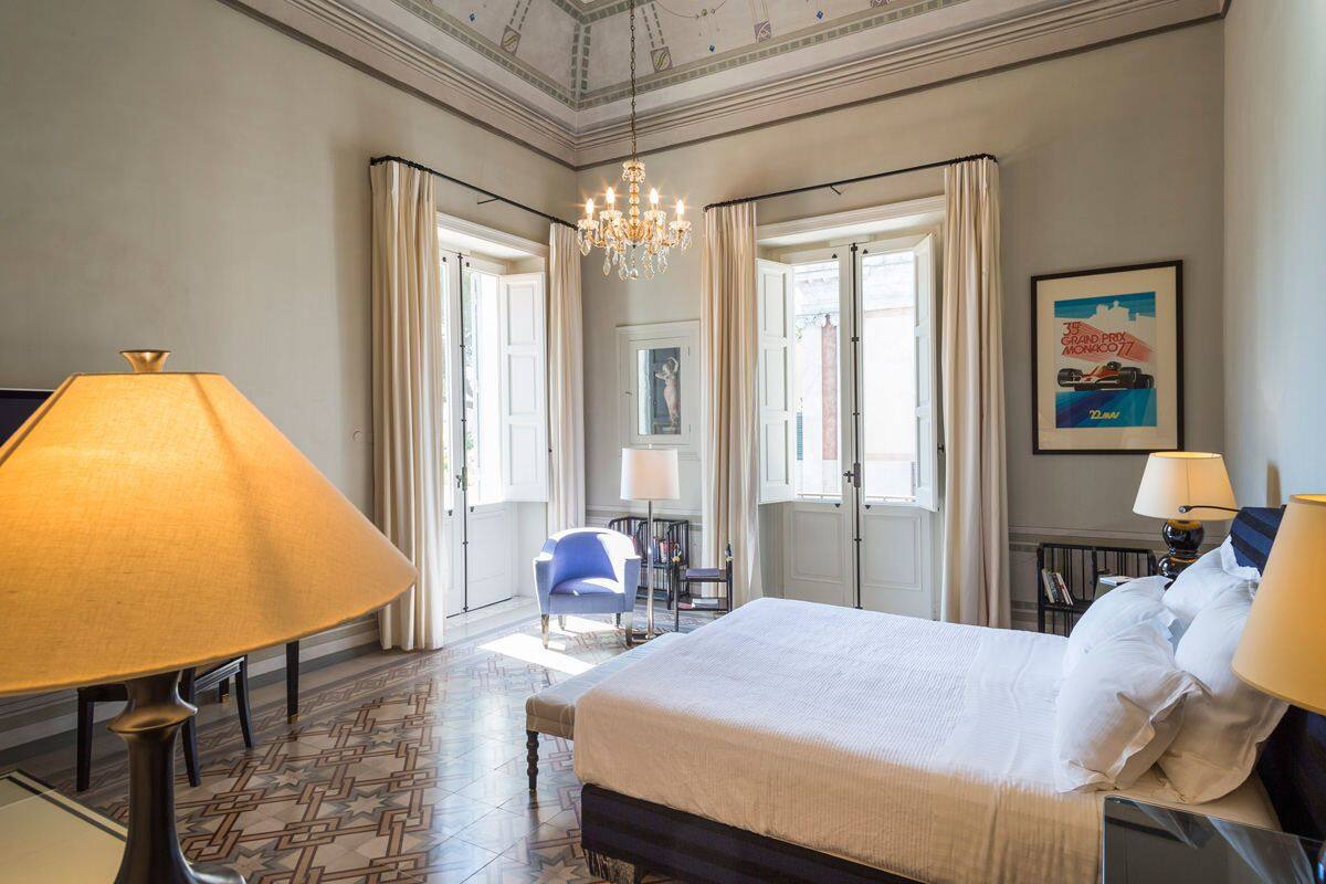 A hotel room with a large bed, two bedside lamps, a chandelier, a desk with a chair, and a framed picture on the wall.