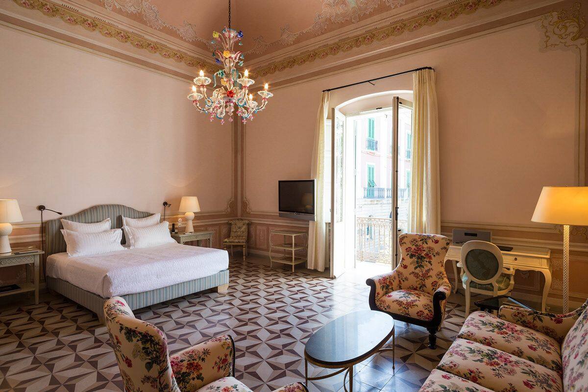 A hotel room with a bed, a desk, a chair, a sofa, a chandelier, and a television.