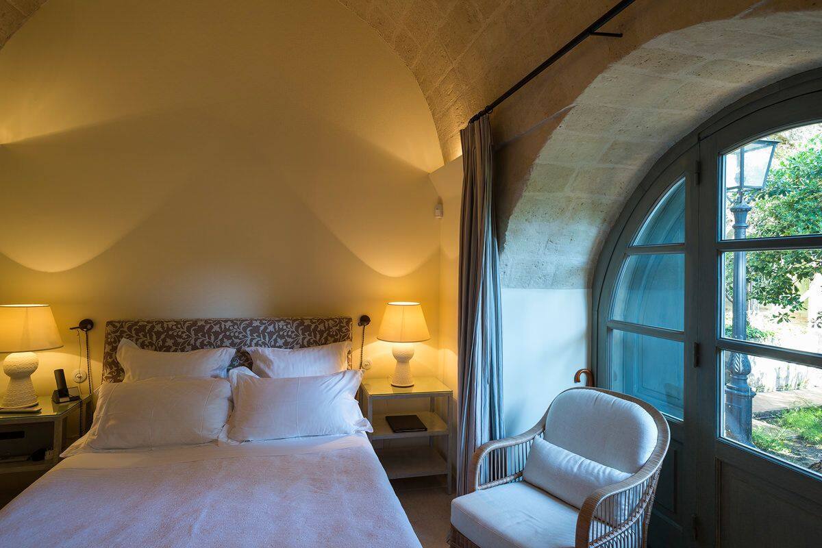 A bedroom with a large bed, two bedside lamps, a nightstand, a chair, and a window with a view of greenery.