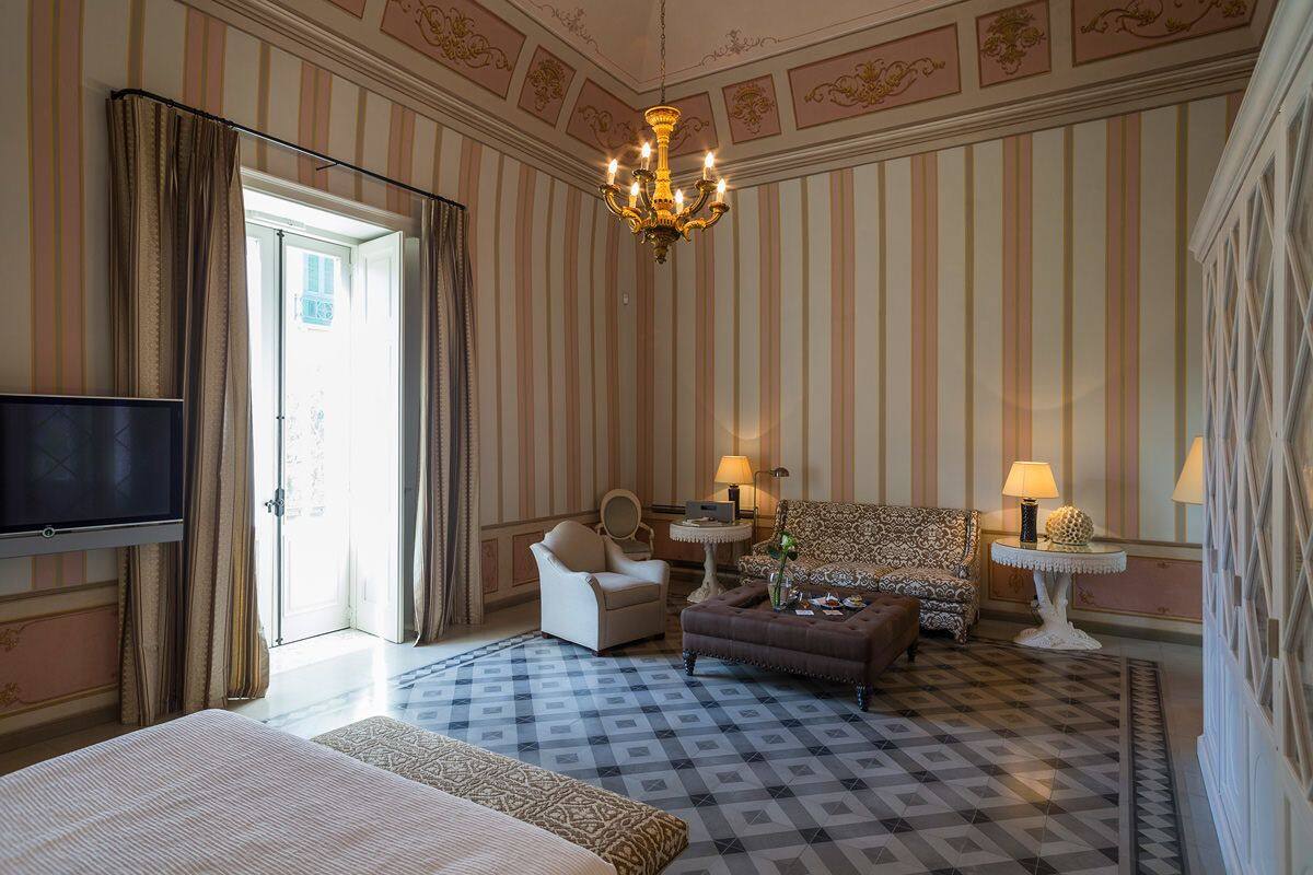 A hotel room with a large bed, a sofa, a chair, a TV, a chandelier, and a patterned floor.