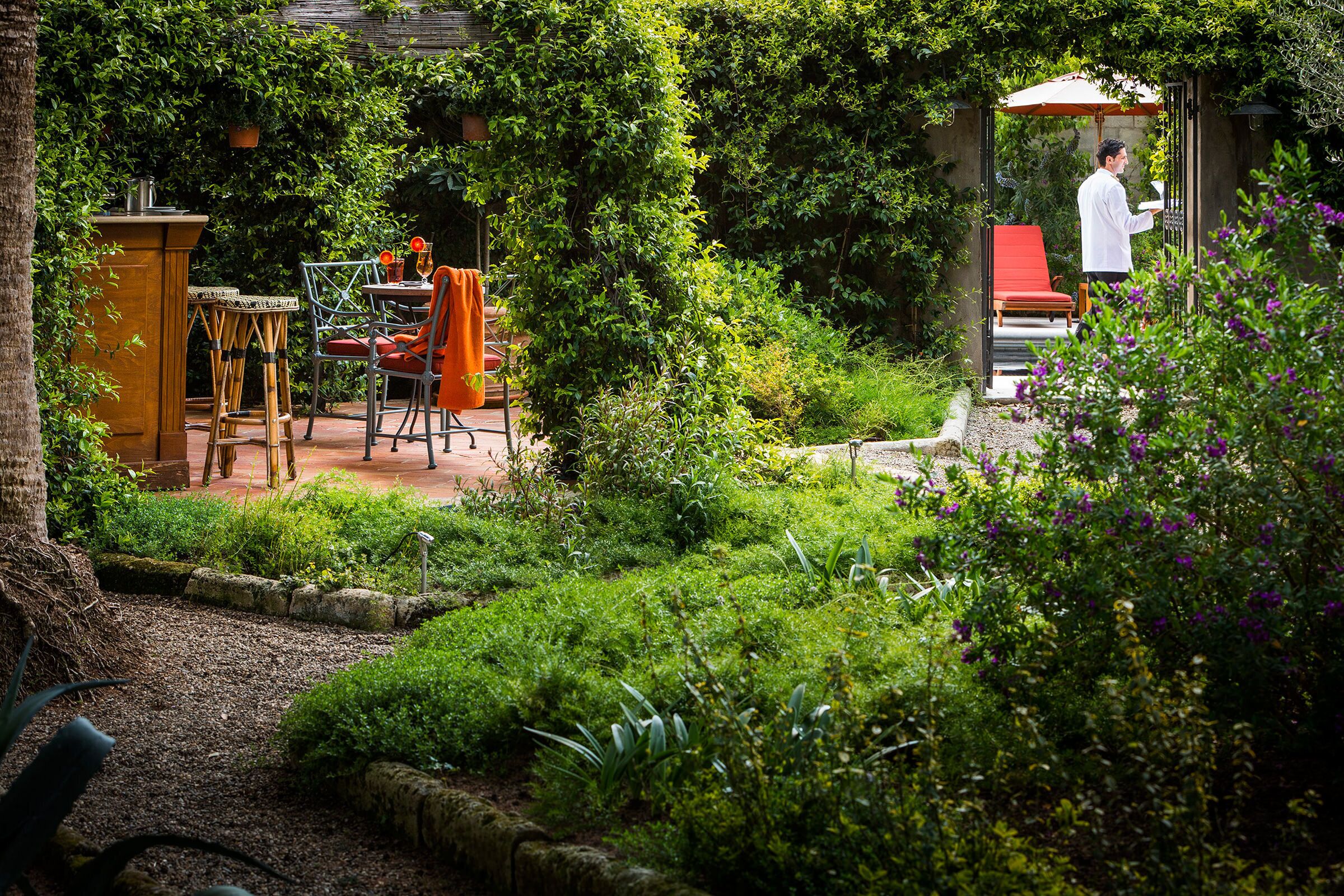Ein üppiger Garten mit einer Terrasse, die einen Tisch, Stühle, einen roten Liegestuhl und eine kleine Bar umfasst.