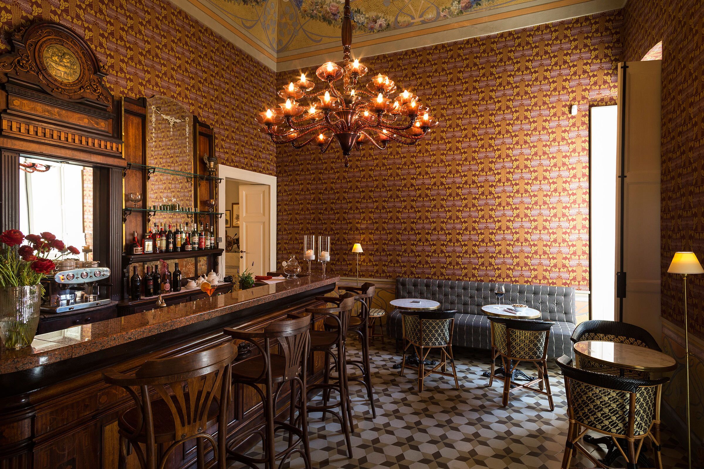 Un’area bar con carta da parati decorata, un lampadario, sgabelli da bar e una zona salotto con divano e poltrone con motivi decorativi.