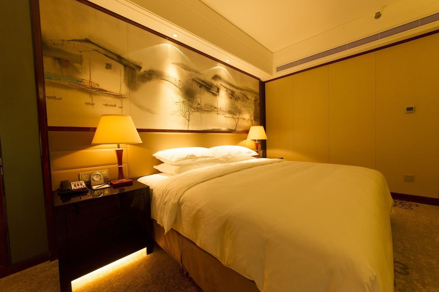A hotel room with a large bed, two bedside lamps, a nightstand with a clock and a telephone, and a wall with a painting.