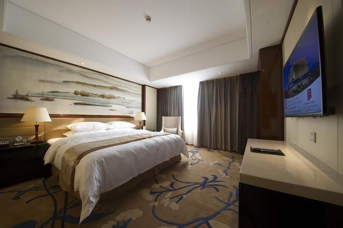 A hotel room with a large bed, a desk with a lamp, a painting on the wall, and a television.