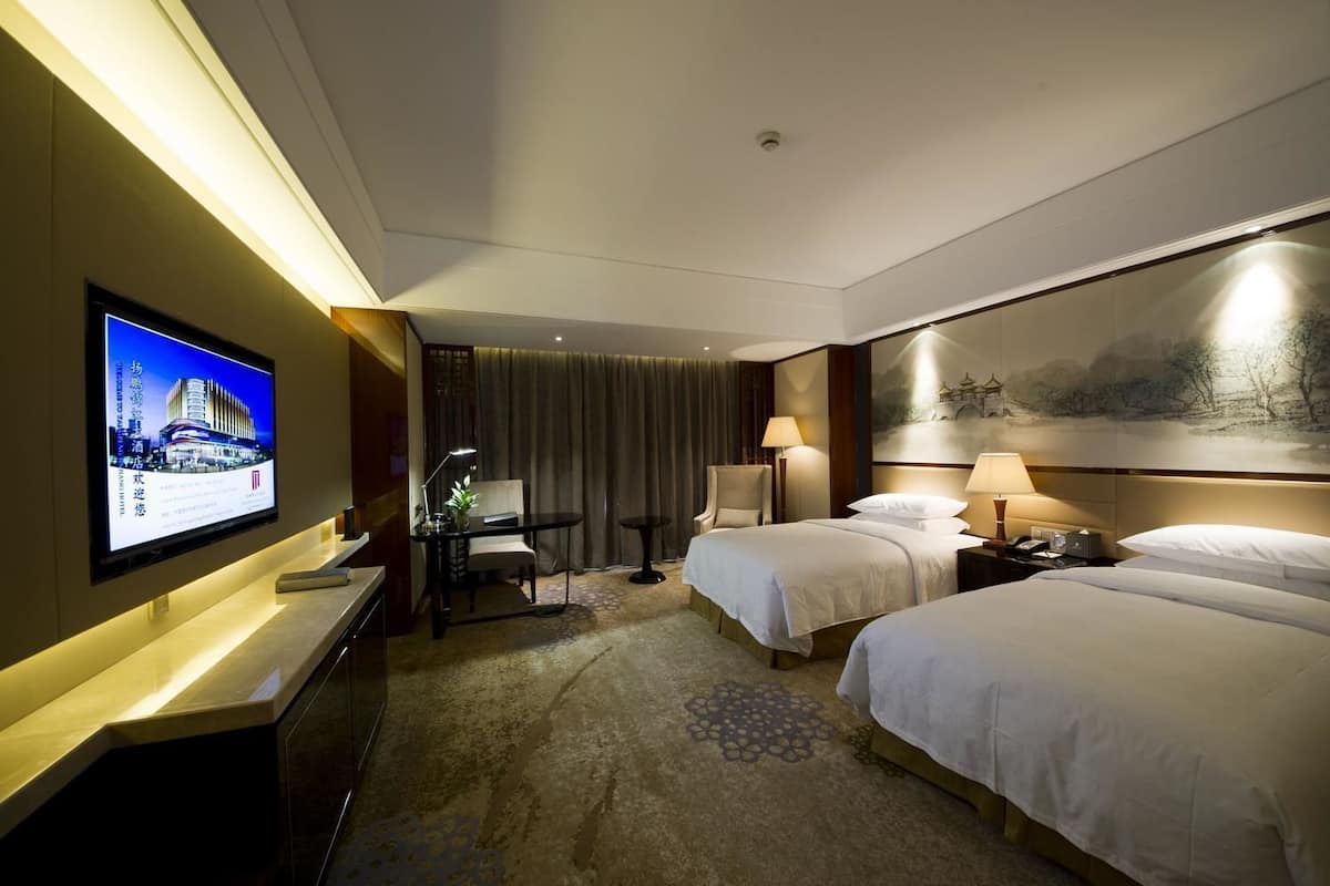 A hotel room with two beds, a desk with a chair, a television, and a painting on the wall.