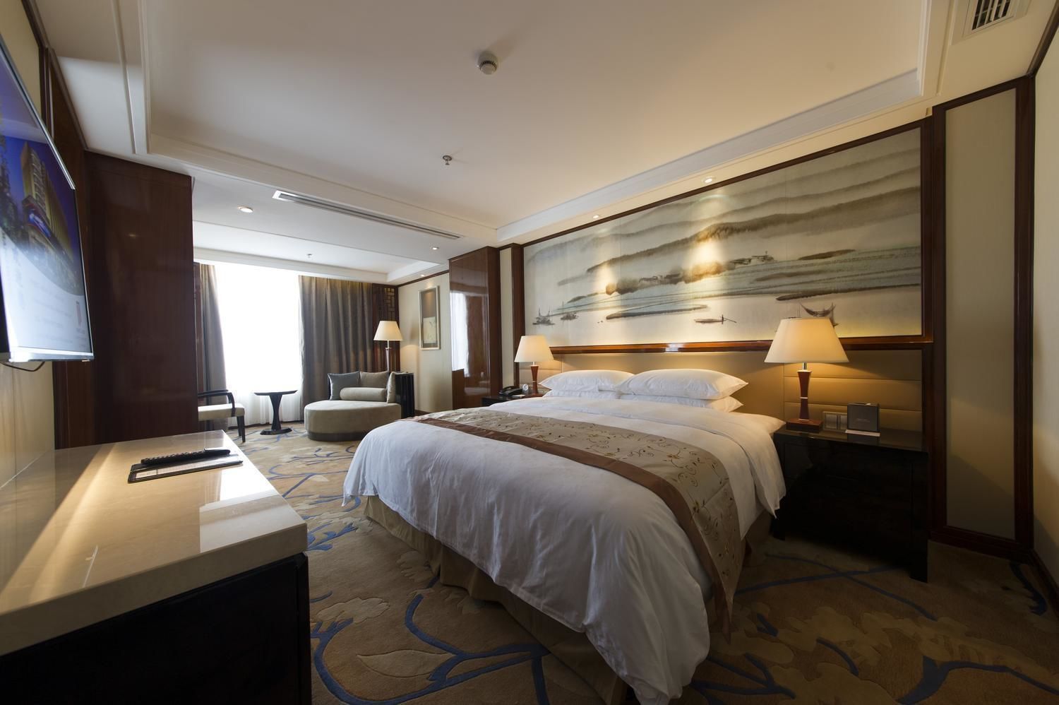 A hotel room with a large bed, a desk with a TV, a chair, and a painting on the wall.