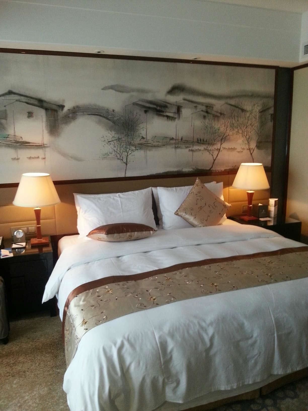 A hotel room with a large bed, two bedside lamps, a nightstand with a clock, and a painting on the wall depicting a landscape.