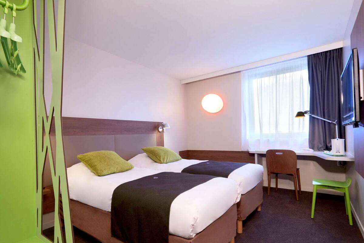 Une chambre d’hôtel avec deux lits, un bureau, une chaise, une télévision et une fenêtre avec des rideaux.