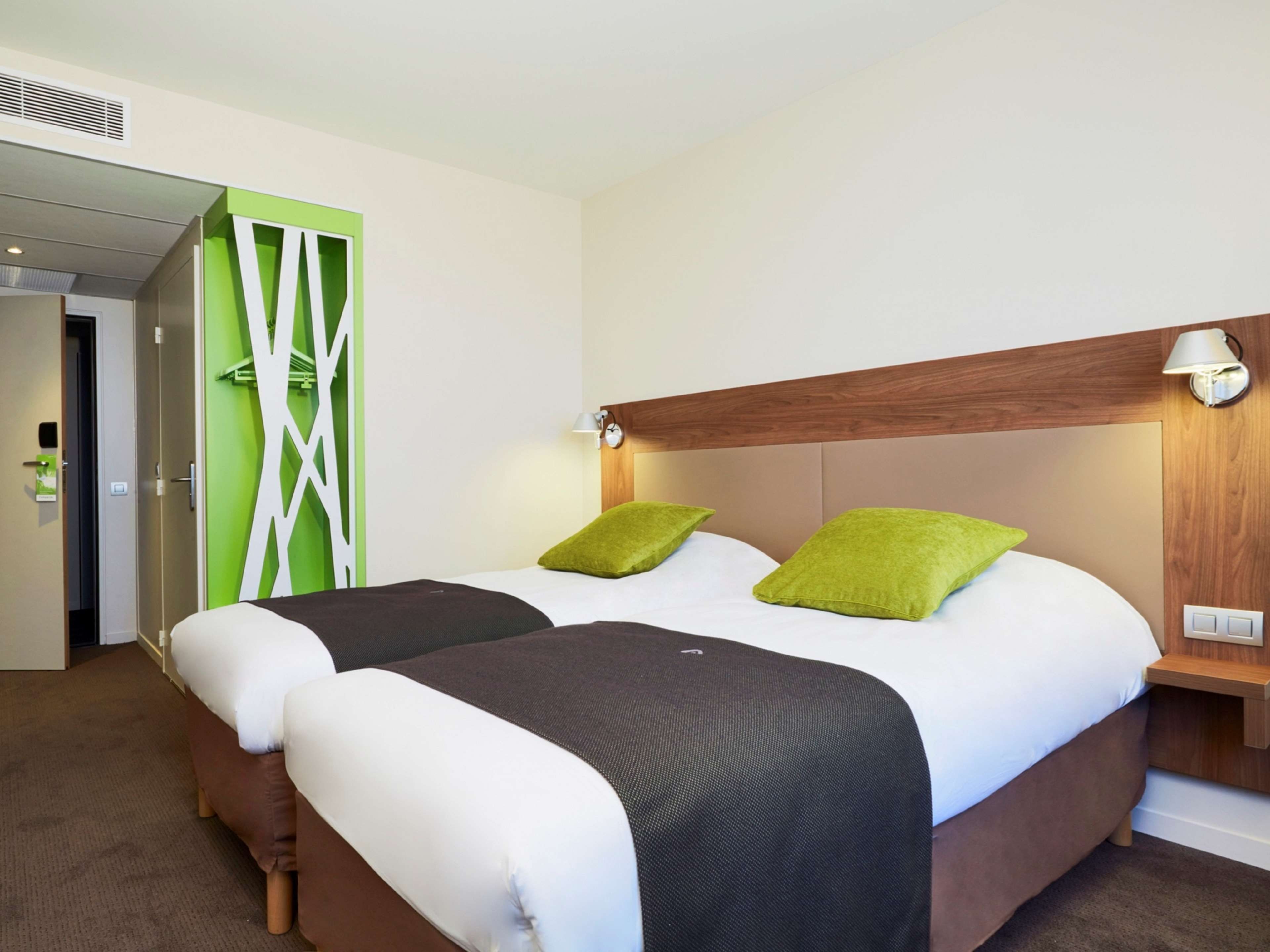 A hotel room with a double bed, two pillows, a bedside table with a lamp, and a green accent wall.