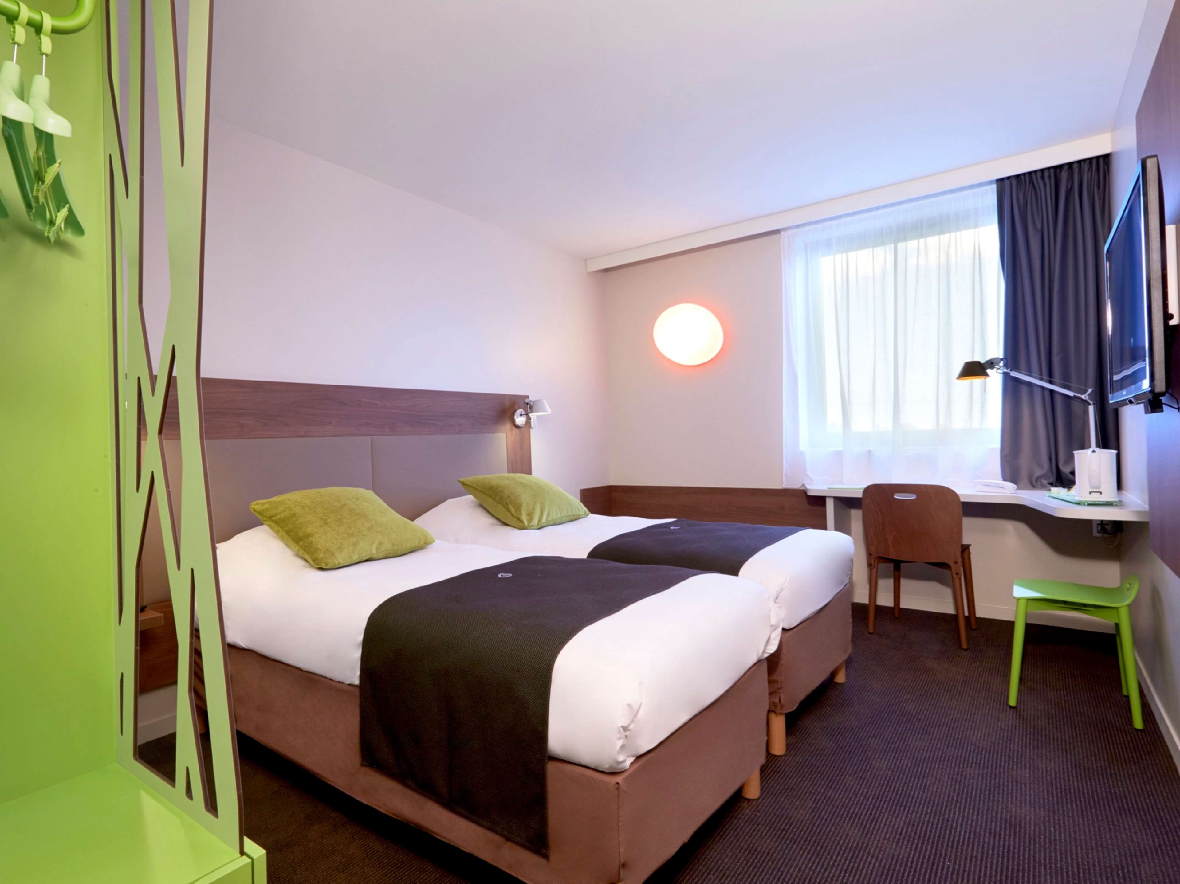 Ein Hotelzimmer mit zwei Betten, einem Schreibtisch, einem Stuhl, einem Fernseher und einem Fenster mit Vorhängen.