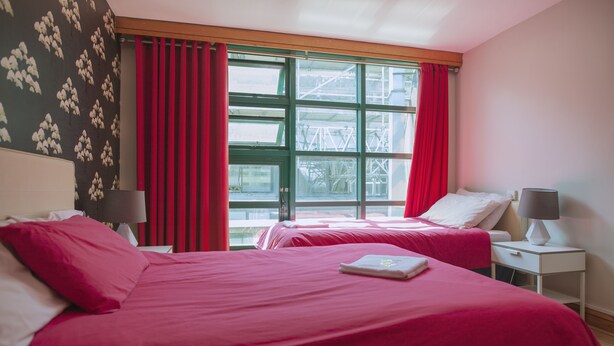 Une chambre d’hôtel avec deux lits, une literie rouge, une fenêtre avec des rideaux rouges et une vue sur une zone industrielle.