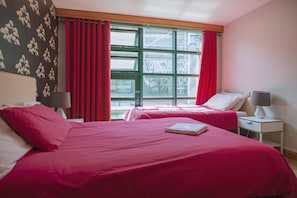 A hotel room with two beds, red bedding, a window with red curtains, and a view of an industrial area. - Abigails Hostel (Dublin)