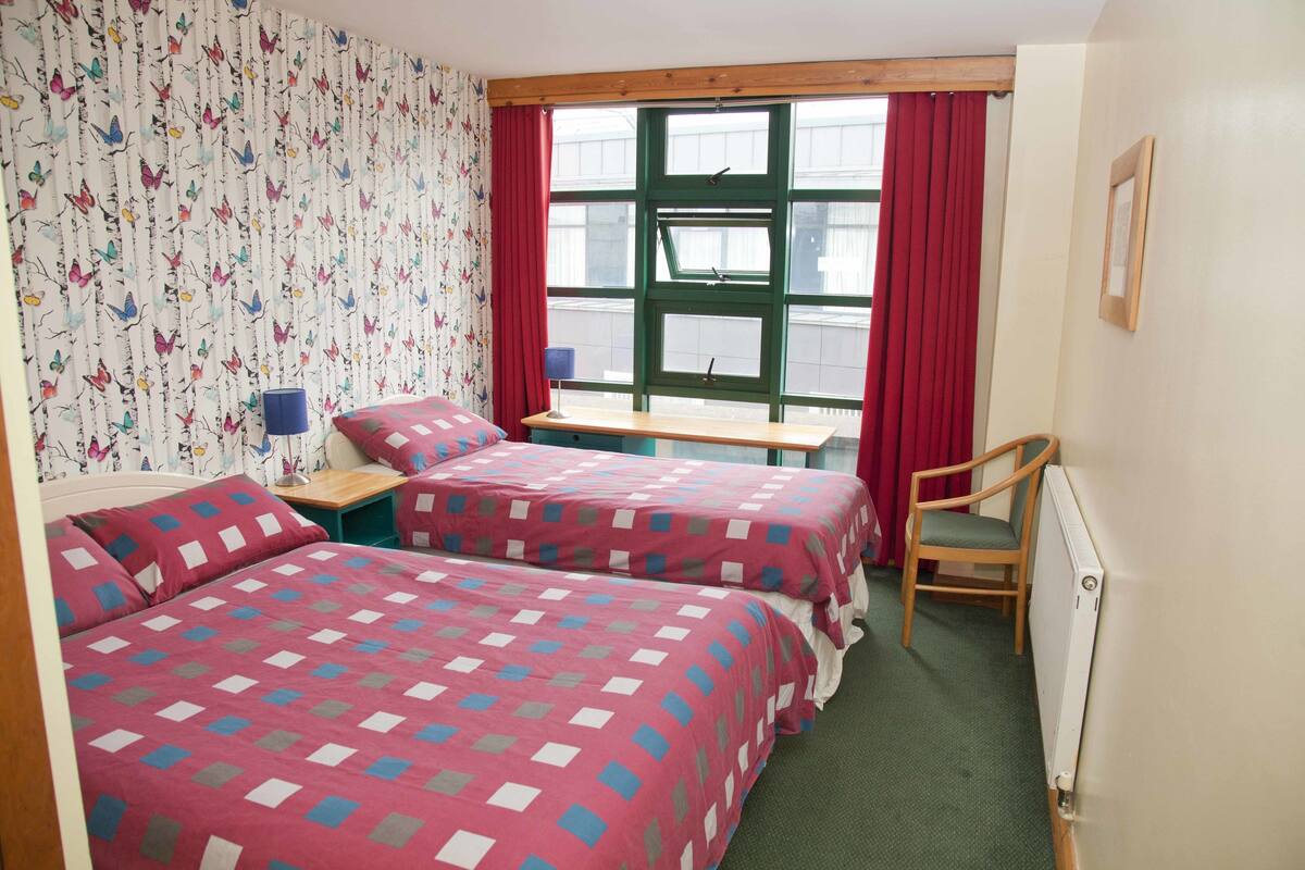 A room with two beds, a desk, a chair, a window with red curtains, and a radiator.