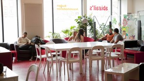 Un groupe de personnes est assis autour d’une table blanche dans une pièce moderne dotée de grandes fenêtres, d’un sol rouge et de diverses plantes.