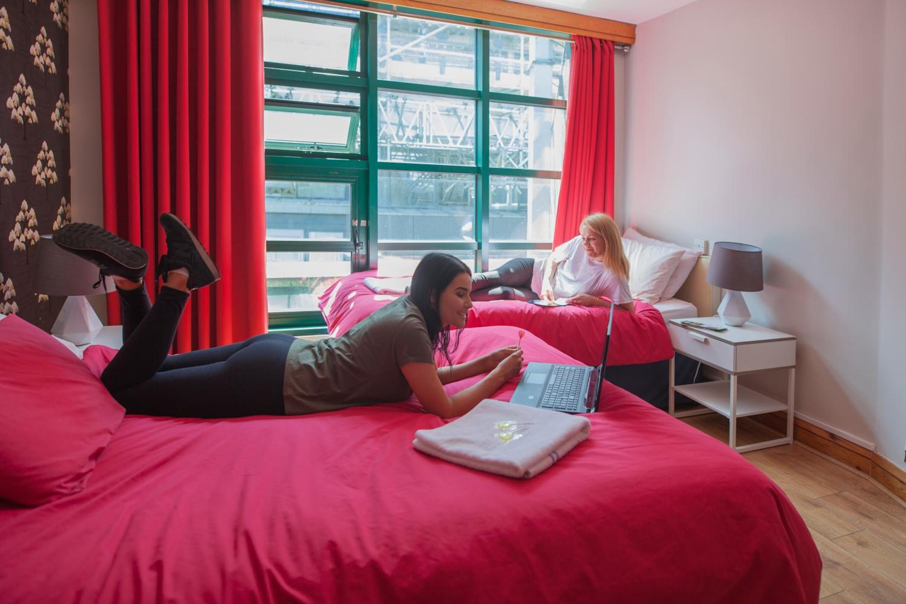 Una habitación de hotel con una cama grande, una computadora portátil y una ventana con cortinas rojas.
