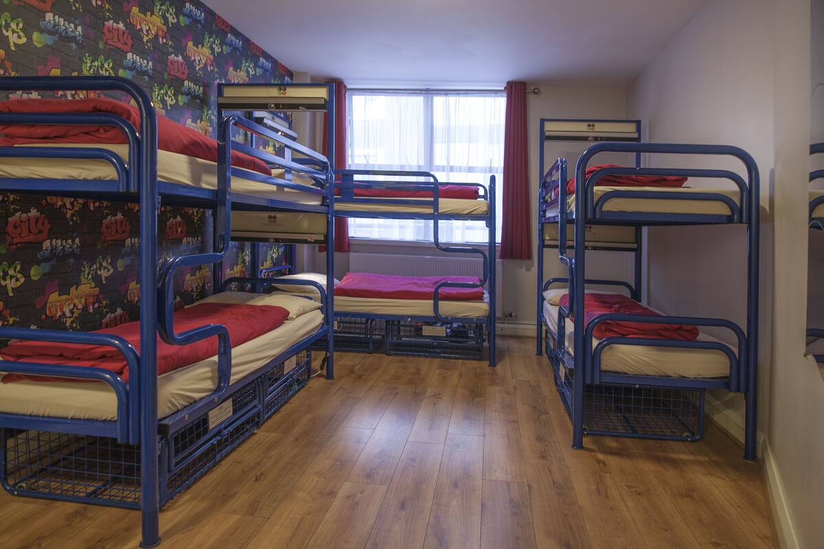 A dormitory room with bunk beds, wooden flooring, and a window with curtains.