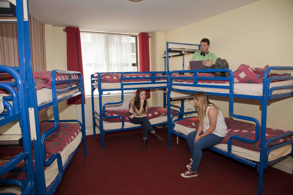 A dormitory room with bunk beds, a window with curtains, and a person using a laptop.