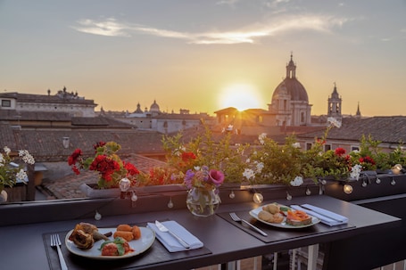 A dining table with plates of food, a vase with flowers, and a view of a cityscape with a prominent dome and bell tower at sunset.