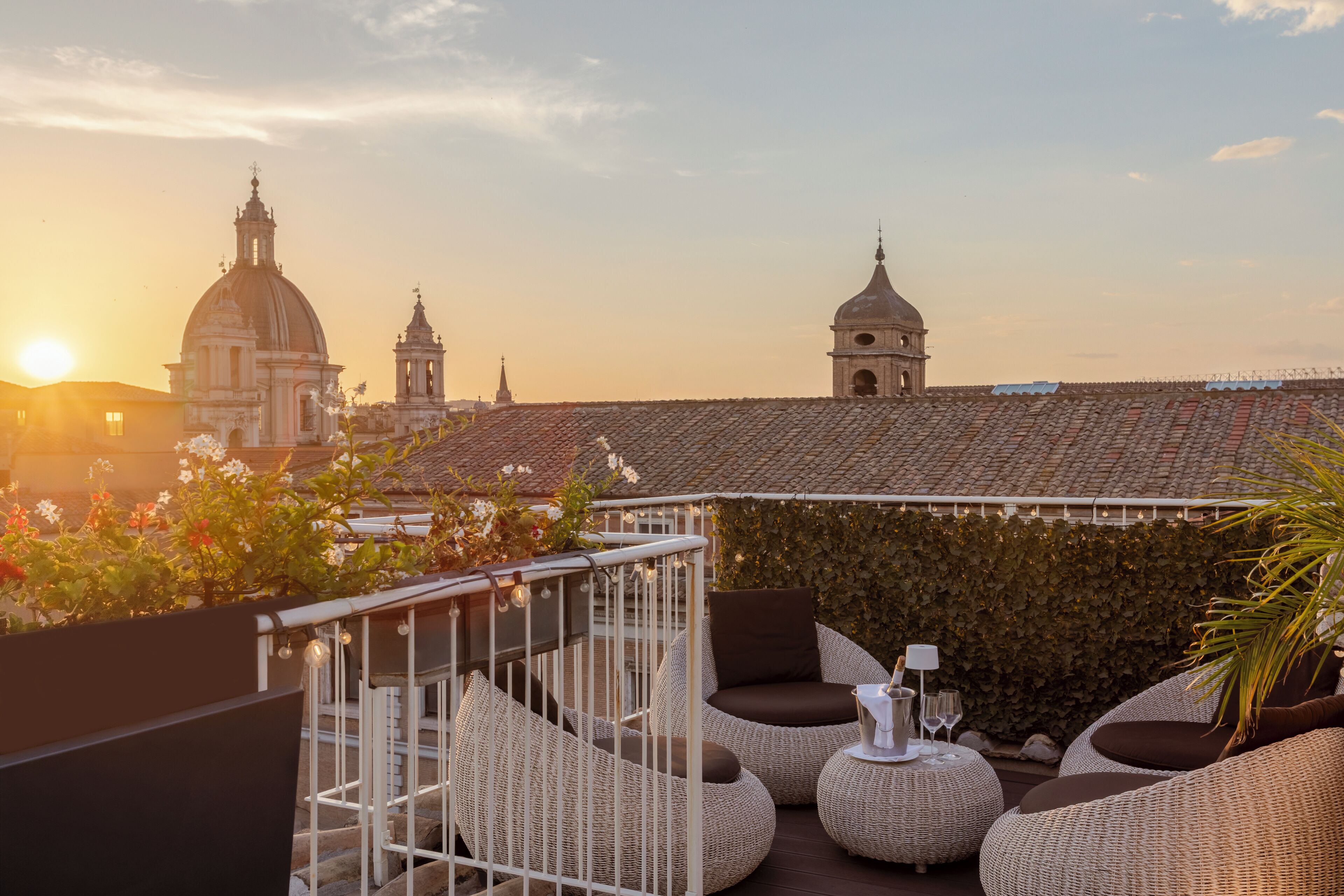 Une terrasse sur le toit, meublée de mobilier en osier, offrant une vue sur des bâtiments historiques et un magnifique coucher de soleil.