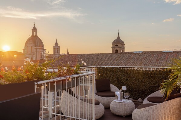 A rooftop terrace with wicker furniture, a view of historic buildings, and a sunset.