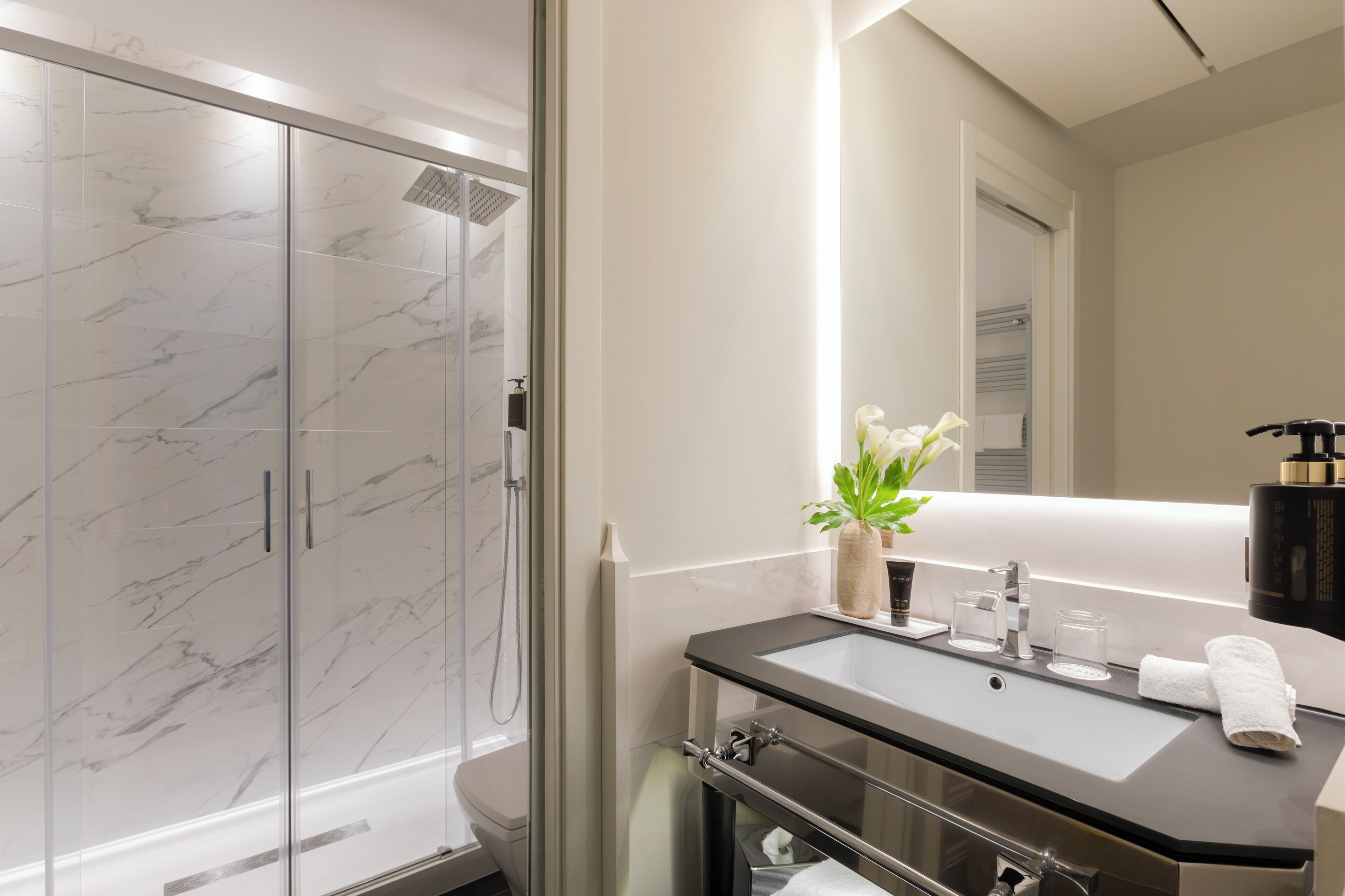 A modern bathroom with a glass shower enclosure, a marble-tiled wall, a sink with a mirror, and a vase with flowers.