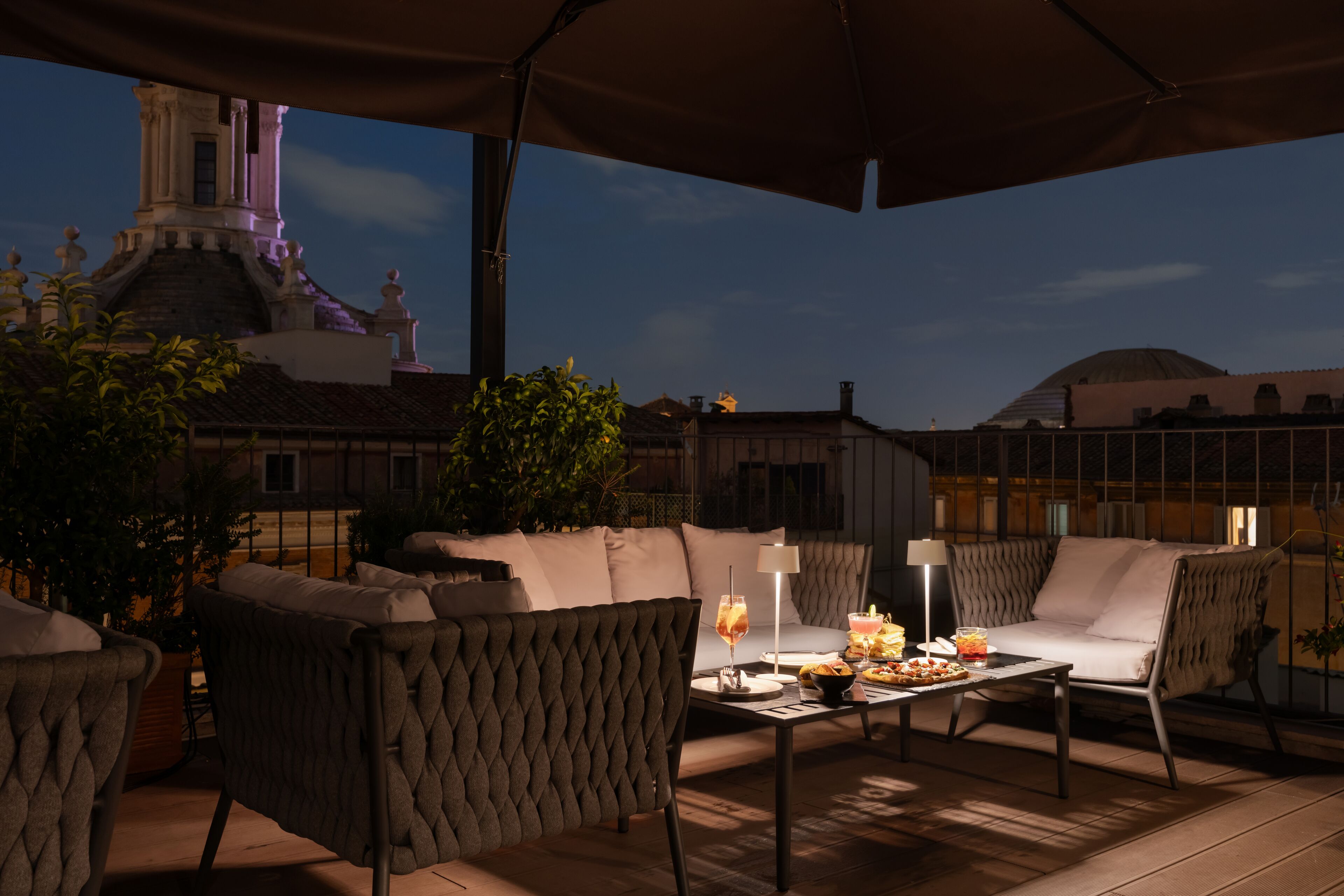 Un patio al aire libre con asientos acolchados, una mesa con comida y bebidas, y vistas a un edificio histórico con una torre.