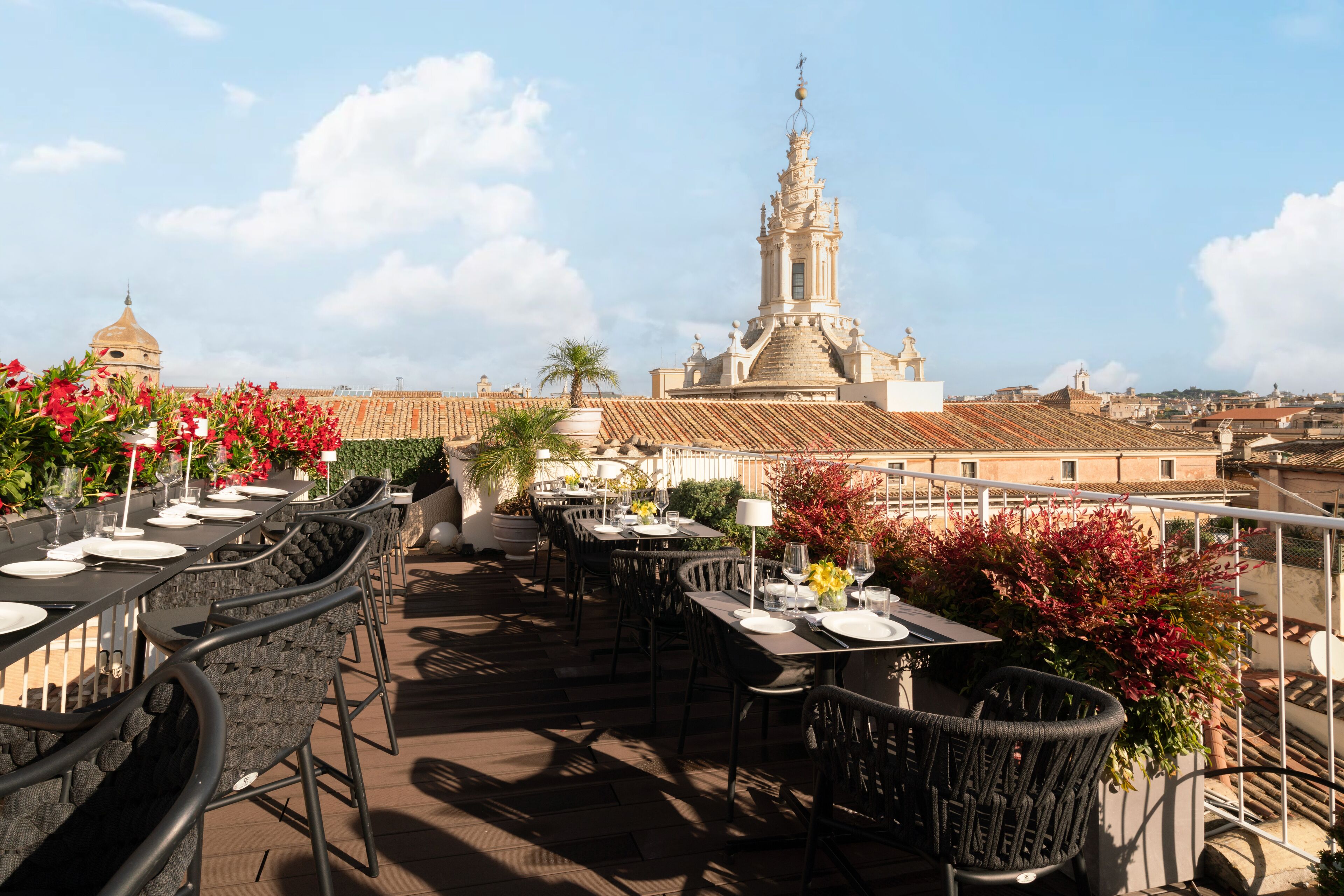 An outdoor dining area with tables and chairs, potted plants, and a view of historic buildings in the background.