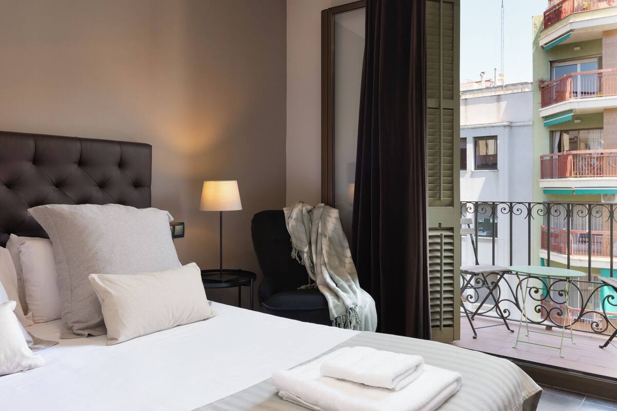 Quarto de hotel com cama, almofadas, uma cadeira, mesa, luminária e vista para a varanda.