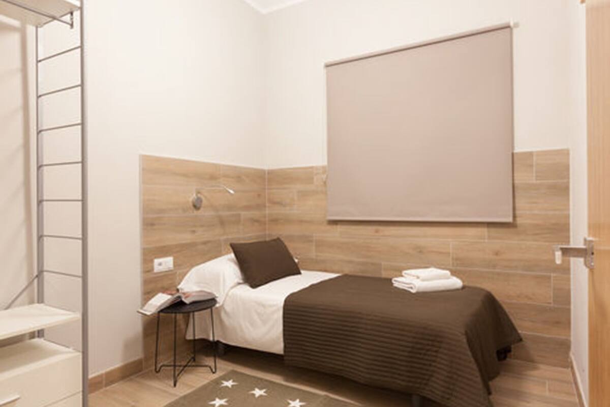 Quarto pequeno e bem organizado com cama, uma cadeira e uma mesinha.
