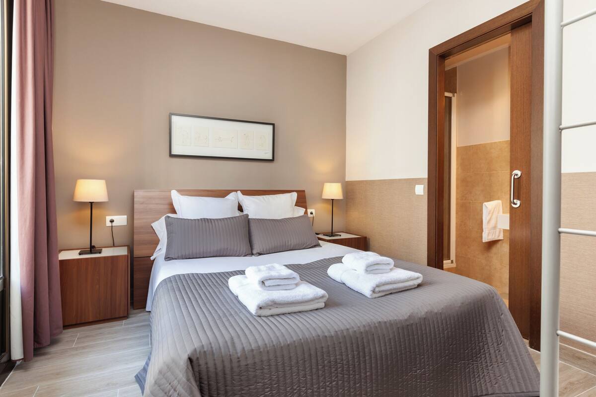 Quarto de hotel com cama, duas mesas de cabeceira com luminárias, um quadro na parede e uma porta com porta-toalhas.