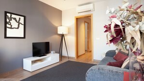 A modern living room with a flat-screen TV, white entertainment unit, floor lamp, grey sofa, and a decorative vase with dried flowers. - Easy Sleep Eixample  (Barcelona)