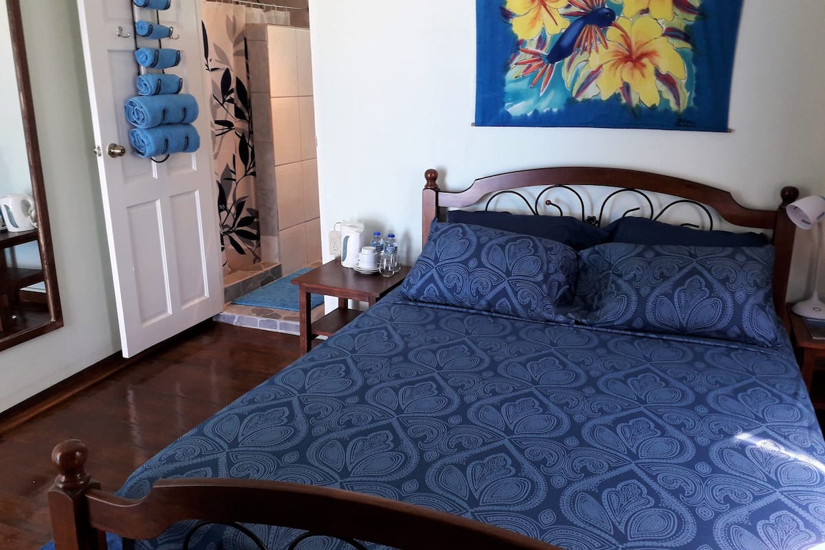 A bedroom with a bed, a nightstand, a painting, a door with towels, and a shower area.