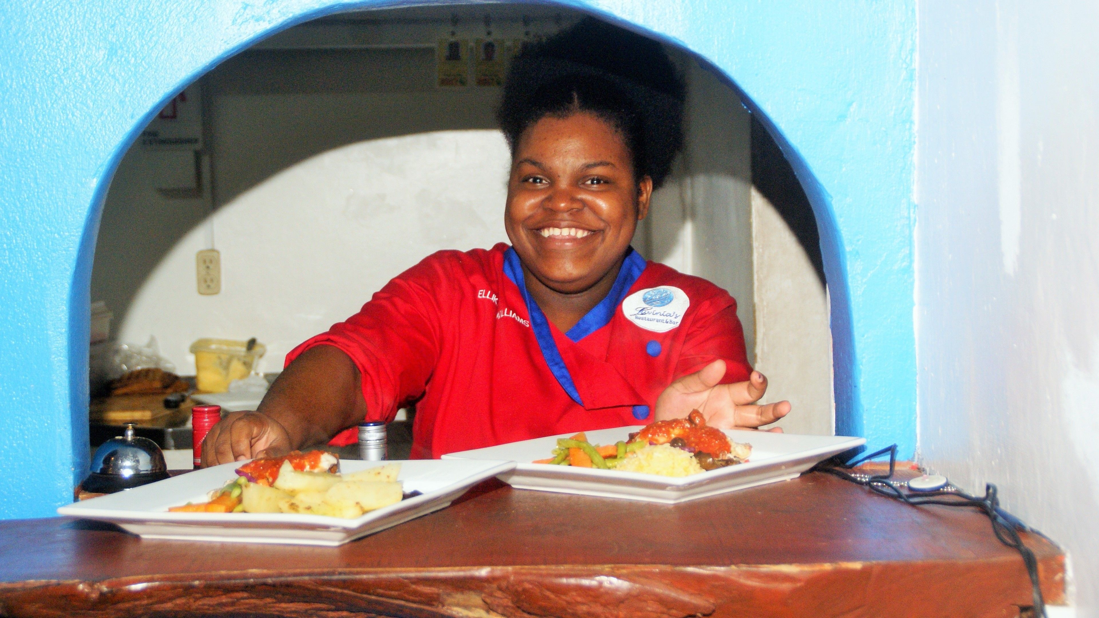 A person in a red uniform serving food from a counter with a blue archway in the background.