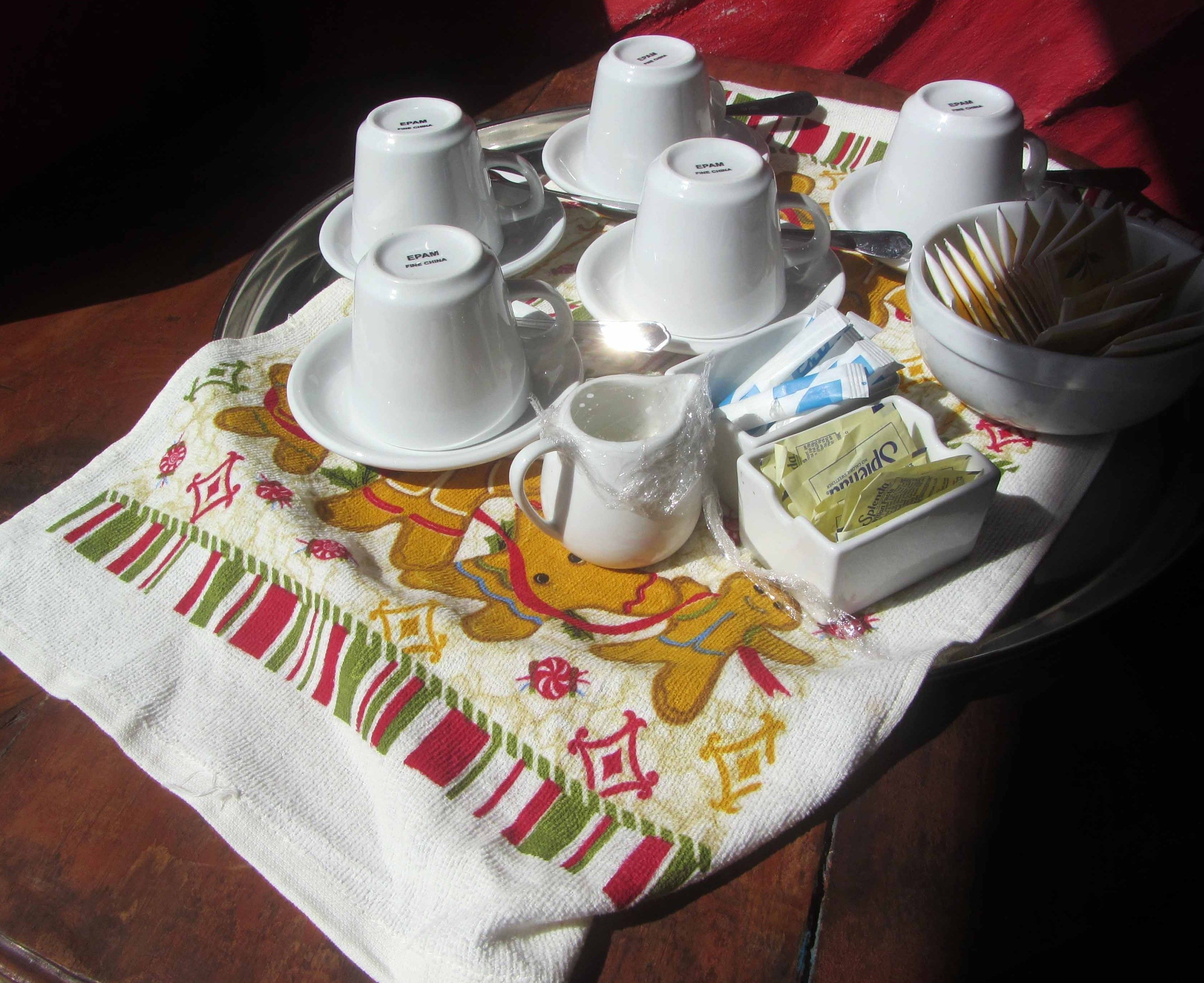Una bandeja con tazas blancas, platillos, una jarrón y una caja de pañuelos sobre un mantel con un diseño festivo.