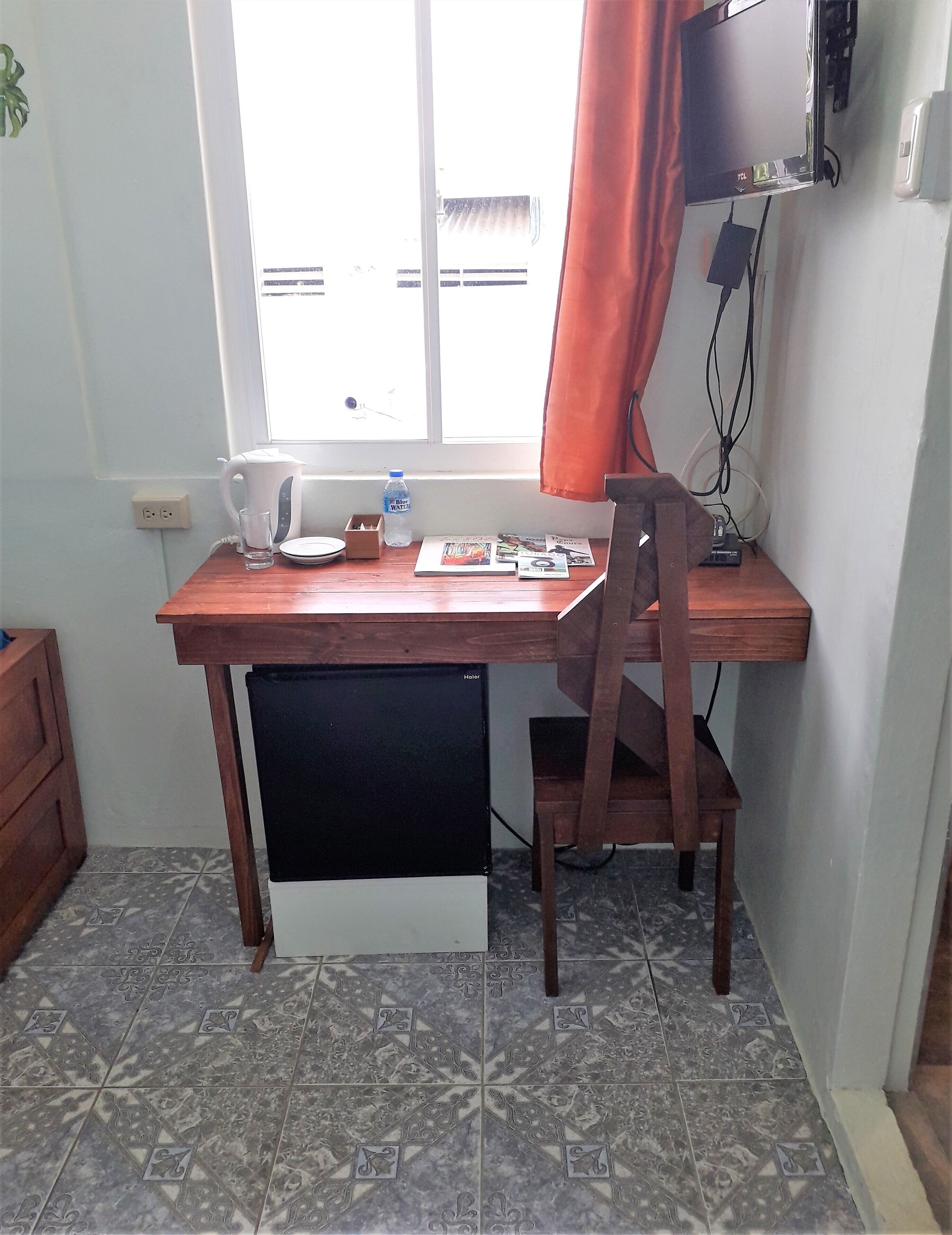 A small, well-lit room with a wooden desk, a chair, a kettle, a glass, and a water bottle.
