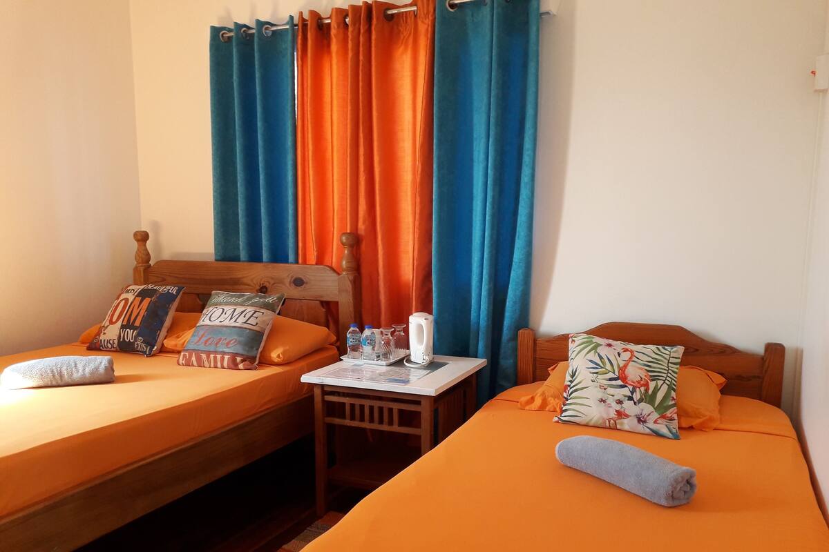 A room with two beds, each with a wooden headboard, orange bedding, and decorative pillows. A small table with a kettle and water bottles is between the beds.