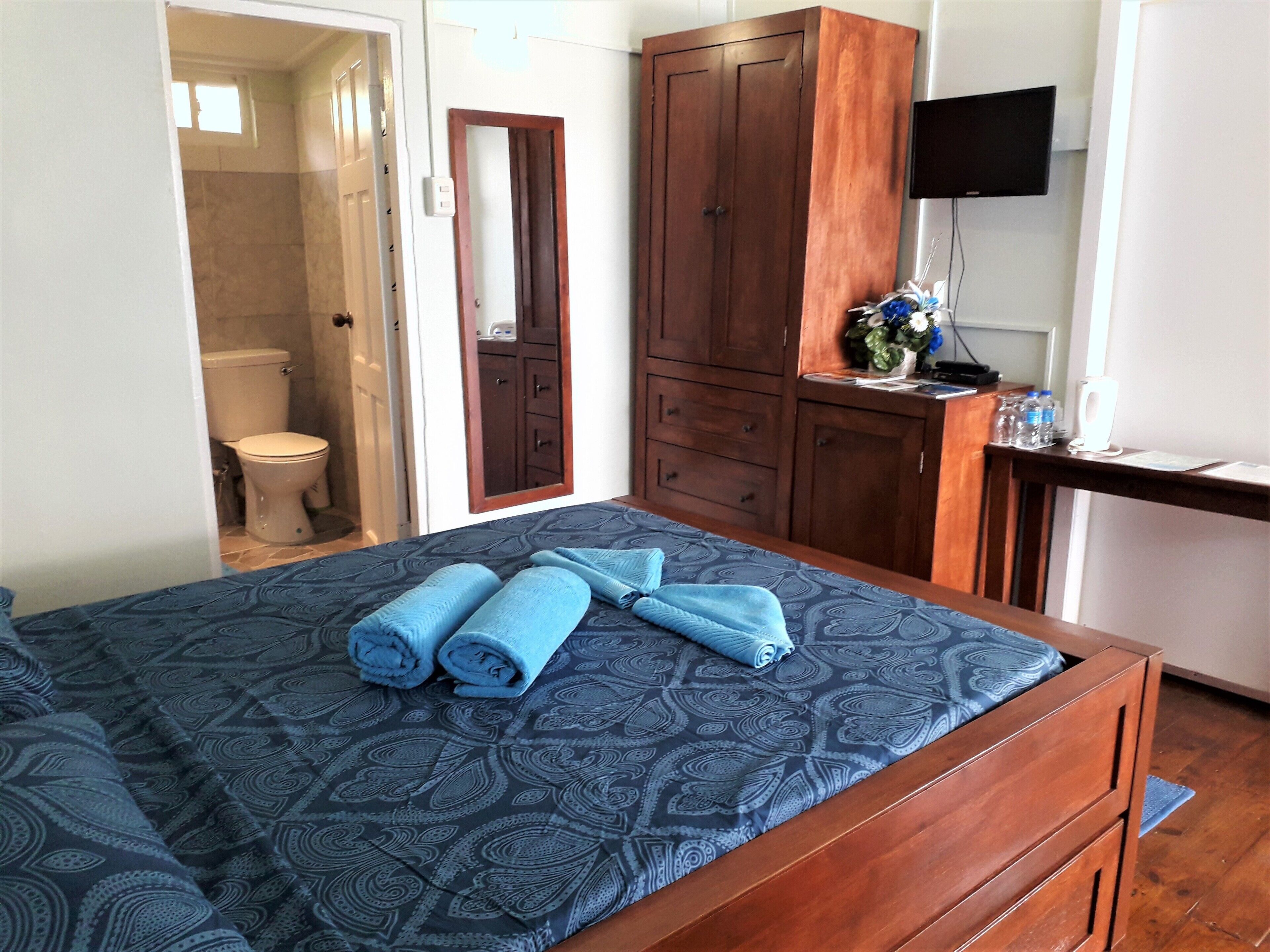 Un dormitorio con cama, armario de madera, escritorio con televisor y baño visible a través de una puerta abierta.
