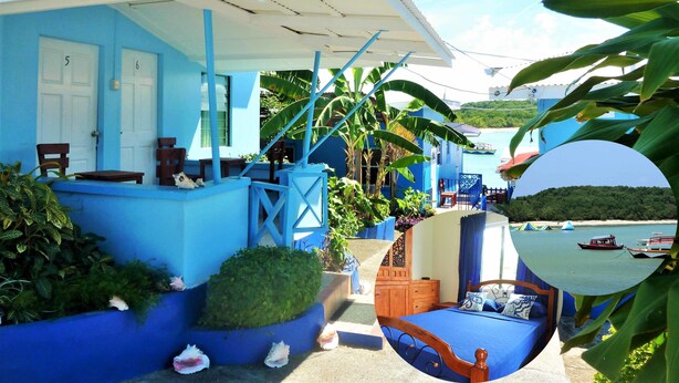 A blue building with numbered doors, a covered outdoor seating area, and a view of boats on the water. - Miller's Guest House (Buccoo)